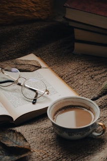 A cozy setting featuring an open book with reading glasses resting on its pages. A steaming cup of coffee is placed nearby on a textured blanket. The scene includes a few scattered leaves and a stack of closed books in the background.
