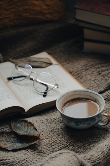 A cozy setting featuring an open book with glasses resting on it, accompanied by a steaming cup of coffee. Nearby are two dried leaves, all placed on a textured, warm-toned fabric surface. A stack of books is partially visible in the background.