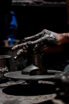 Hands covered in clay are intricately shaping a pottery piece on a rotating potter's wheel. The environment is dimly lit, highlighting the texture and detail of both the hands and the clay. The focus is on the creative process and craftsmanship involved in pottery making.