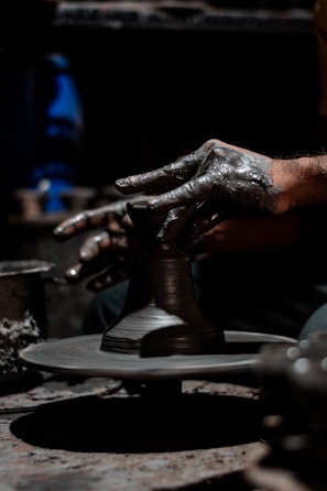 Hands covered in clay are intricately shaping a pottery piece on a rotating potter's wheel. The environment is dimly lit, highlighting the texture and detail of both the hands and the clay. The focus is on the creative process and craftsmanship involved in pottery making.