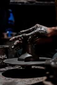 Hands covered in clay are intricately shaping a pottery piece on a rotating potter's wheel. The environment is dimly lit, highlighting the texture and detail of both the hands and the clay. The focus is on the creative process and craftsmanship involved in pottery making.