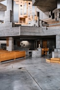 A spacious, modern interior features extensive use of concrete and wood. The architecture displays a series of geometric shapes with stairs and platforms. A large, intricate organ pipes structure is prominently situated against a backdrop of gray concrete. The space has wooden benches, suggesting it may be a church or auditorium.