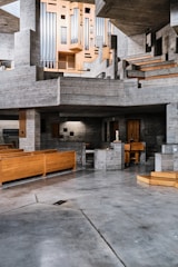 A spacious, modern interior features extensive use of concrete and wood. The architecture displays a series of geometric shapes with stairs and platforms. A large, intricate organ pipes structure is prominently situated against a backdrop of gray concrete. The space has wooden benches, suggesting it may be a church or auditorium.