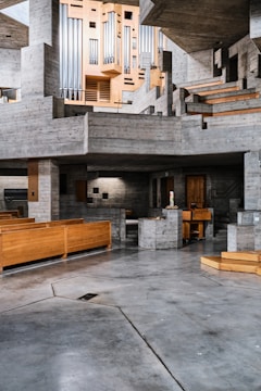A spacious, modern interior features extensive use of concrete and wood. The architecture displays a series of geometric shapes with stairs and platforms. A large, intricate organ pipes structure is prominently situated against a backdrop of gray concrete. The space has wooden benches, suggesting it may be a church or auditorium.