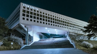 A modern building with a unique architectural design, prominently displaying the name 'Star Center' on its facade. The structure is elevated, supported by pillars, and features a wide staircase leading up to the entrance. The surrounding area is landscaped with bushes and trees. The scene is set at night, and the building is illuminated by exterior lights.