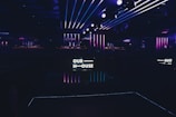 A dimly lit interior space with neon lights and a sign that reads 'OUR HOUSE,' featuring vibrant purple and blue stripes along the walls and ceiling. The atmosphere suggests a nightclub or contemporary art installation.