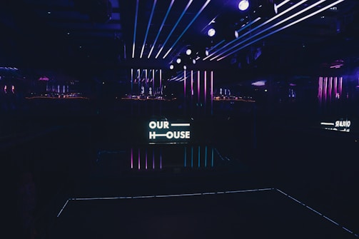 A dimly lit interior space with neon lights and a sign that reads 'OUR HOUSE,' featuring vibrant purple and blue stripes along the walls and ceiling. The atmosphere suggests a nightclub or contemporary art installation.