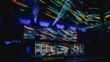 A vibrant nightclub interior featuring a visually striking light display with colorful neon streaks across the walls and ceiling. The design creates an energetic and dynamic atmosphere, enhanced by multiple beams of light. Speaker arrays are visible, suggesting a lively music scene.