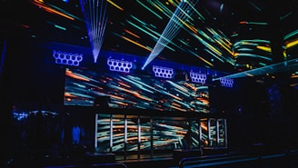 A vibrant nightclub interior featuring a visually striking light display with colorful neon streaks across the walls and ceiling. The design creates an energetic and dynamic atmosphere, enhanced by multiple beams of light. Speaker arrays are visible, suggesting a lively music scene.