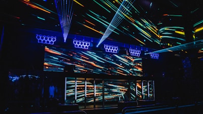 A vibrant nightclub interior featuring a visually striking light display with colorful neon streaks across the walls and ceiling. The design creates an energetic and dynamic atmosphere, enhanced by multiple beams of light. Speaker arrays are visible, suggesting a lively music scene.