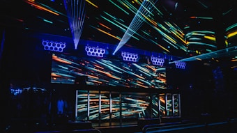 A vibrant nightclub interior featuring a visually striking light display with colorful neon streaks across the walls and ceiling. The design creates an energetic and dynamic atmosphere, enhanced by multiple beams of light. Speaker arrays are visible, suggesting a lively music scene.