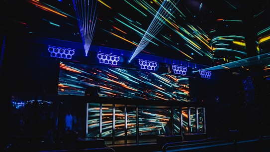 A vibrant nightclub interior featuring a visually striking light display with colorful neon streaks across the walls and ceiling. The design creates an energetic and dynamic atmosphere, enhanced by multiple beams of light. Speaker arrays are visible, suggesting a lively music scene.