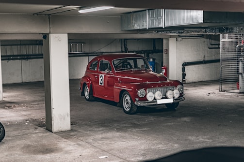 A vintage red car with the number three displayed on its side is parked in an underground garage. The car features multiple auxiliary lights on the front bumper and a classic design typical of mid-20th-century vehicles. The garage has a concrete floor and ceiling, with visible pillars and some industrial elements like pipes and a metal cage in the background.