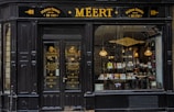 An old-fashioned storefront with 'MEERT' written prominently at the top, indicating a traditional confectionery shop. The signage indicates the shop was established in 1761. The entrance features double doors with gold lettering advertising various sweets and biscuits. Through the large window display, various candies and chocolates are visible inside, illuminated by warm hanging lights.