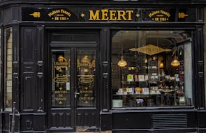 An old-fashioned storefront with 'MEERT' written prominently at the top, indicating a traditional confectionery shop. The signage indicates the shop was established in 1761. The entrance features double doors with gold lettering advertising various sweets and biscuits. Through the large window display, various candies and chocolates are visible inside, illuminated by warm hanging lights.