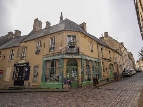 A quaint, rustic street corner features an old building housing a restaurant with green accents and large windows. The structure has a stone facade with several chimneys and a steeply pitched roof. Cobbled streets and sidewalks add to the charm, with several decorative signs and menu boards outside the restaurant.