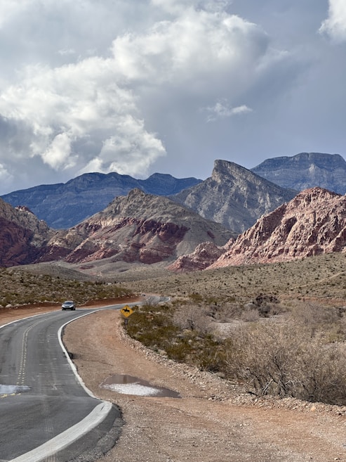 A winding road stretches through a desert landscape with rugged mountains in the background. The terrain is mostly barren with some vegetation. A car is traveling on the road, and a road sign is visible. The sky is overcast with large, puffy clouds.