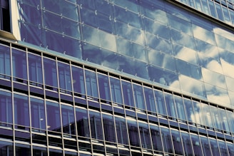 The image features a modern glass building reflecting the sky with clouds. The architecture displays clean lines and a structured grid pattern formed by the window frames.