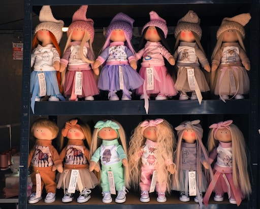 On display are two rows of handcrafted dolls in a store, each dressed in colorful outfits with hats. The top row features six dolls in pastel dresses and knitted hats, while the dolls on the bottom row are dressed in casual outfits with various types of headbands. The dolls have simple facial features with blushed cheeks.
