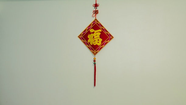 A decorative Chinese hanging ornament with a red diamond-shaped design, featuring a large yellow Chinese character in the center. Intricate patterns in gold adorn the borders. It is suspended from a wall with red and gold tassels and beads hanging below.