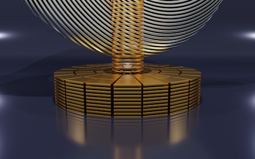 A futuristic, abstract design featuring a series of stacked golden disks forming a circular base. Rising from the center is a column of tightly coiled golden rings, with a large, swirling metallic structure resembling a helix or wave, hovering above. The image has a sleek, polished appearance with reflective surfaces and soft lighting.