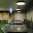 A modern interior setting featuring a series of black tables and grey cushioned chairs. The space is dimly lit by several pendant lights hanging from the ceiling. A neon diamond-shaped light fixture stands out on a textured wall, with plants in large white pots adding greenery. The walls are stylishly decorated with textured patterns, and there are wooden accents and a green floor.