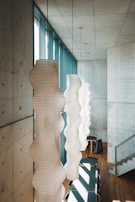 Elegant pendant lights with a wavy, white paper design hang in a modern interior with concrete walls and wooden flooring. Large windows allow natural light to flood the space, creating a calm ambiance. The room is furnished with a row of dark chairs and tables, and a staircase with metal railings is visible in the background.