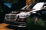 A luxury car with a shiny exterior is parked indoors. The vehicle features sleek lines, a prominent grille with a logo on top, and polished metallic accents. The lighting enhances its glossy surface, emphasizing the car's premium design and elegance. The background is subtly illuminated, with some greenery partially visible.