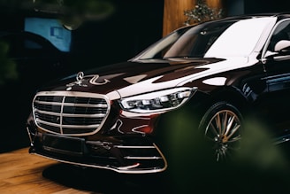 A luxury car with a shiny exterior is parked indoors. The vehicle features sleek lines, a prominent grille with a logo on top, and polished metallic accents. The lighting enhances its glossy surface, emphasizing the car's premium design and elegance. The background is subtly illuminated, with some greenery partially visible.