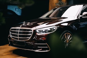 A luxury car with a shiny exterior is parked indoors. The vehicle features sleek lines, a prominent grille with a logo on top, and polished metallic accents. The lighting enhances its glossy surface, emphasizing the car's premium design and elegance. The background is subtly illuminated, with some greenery partially visible.