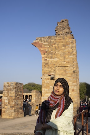 A woman wearing a colorful headscarf and light-colored garment stands in front of ancient stone ruins. The ruins consist of tall, weathered stone structures with intricate brick patterns. The sky is clear and blue, indicating a sunny day. There are several people visible in the background exploring the site.