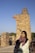 A woman wearing a colorful headscarf and light-colored garment stands in front of ancient stone ruins. The ruins consist of tall, weathered stone structures with intricate brick patterns. The sky is clear and blue, indicating a sunny day. There are several people visible in the background exploring the site.