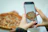Smiling woman enjoying a slice of pizza at home with a smartphone nearby.