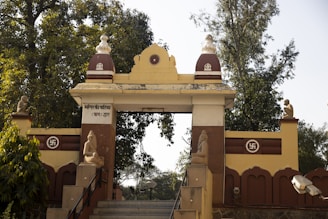 A decorative entrance archway in a temple complex features detailed architectural elements and statues. Swastika symbols are prominently displayed, and there are lush green trees in the background.