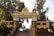 A decorative entrance archway in a temple complex features detailed architectural elements and statues. Swastika symbols are prominently displayed, and there are lush green trees in the background.