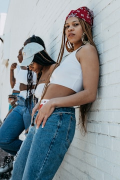 A group of people are leaning against a brick wall, dressed in casual attire with denim jeans and cropped tops. One individual in the foreground wears a red bandana and hoop earrings, while others in the background wear hats and roller skates.