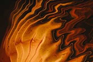 Swirling patterns of black, orange, and amber create a mesmerizing abstract design reminiscent of molten lava or flowing liquid. The smooth, wavy lines mix seamlessly, with darker shades on the edges transitioning to lighter hues in the center. A few small bubbles or specks dot the surface, adding texture to the fluid appearance.
