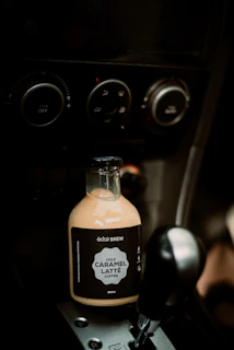 A bottle of cold caramel latte coffee is positioned in the cup holder of a car's interior, surrounded by the controls and dials of a car console.