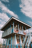 A two-story building with a rustic design, featuring blue and white painted wooden pillars and railings. The house is elevated with an external staircase leading to the second floor, which has wooden railings painted in a combination of red and blue. The exterior includes hanging plants and draped fabric on the upper balcony. The sky above is clear with scattered clouds.