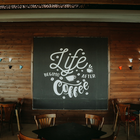 A cozy coffee shop interior features wooden walls and several wooden chairs around dark tables. The focal point is a large chalkboard sign with the phrase 'Life begins after coffee' written in decorative white lettering. Colorful upside-down coffee cups are mounted on the wall as decoration.