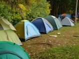 A row of colorful camping tents is set up on a grassy field next to a lush green forest. The tents vary in shades including green, blue, and gray. The area seems organized, and the ground is scattered with some dry leaves and twigs. It is daytime, and the overall environment appears calm and inviting for outdoor activities.