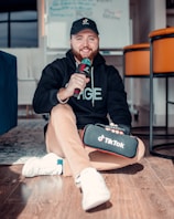 A person wearing a black hoodie and cap is sitting on the floor holding a microphone and a TikTok-branded case. The room has modern furniture, including orange chairs and a blue couch.