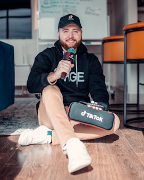 A person wearing a black hoodie and cap is sitting on the floor holding a microphone and a TikTok-branded case. The room has modern furniture, including orange chairs and a blue couch.