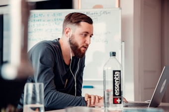 A man with a beard wearing a long-sleeve shirt and earphones is working on a laptop at a desk. A bottle of water labeled 