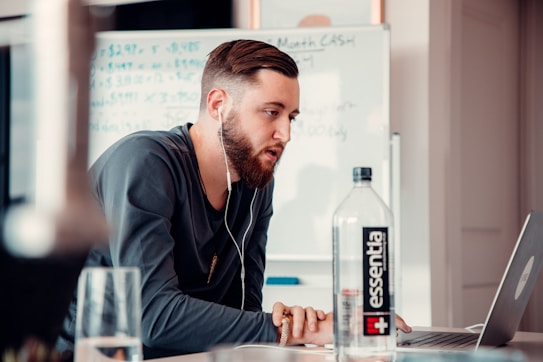 A man with a beard wearing a long-sleeve shirt and earphones is working on a laptop at a desk. A bottle of water labeled 