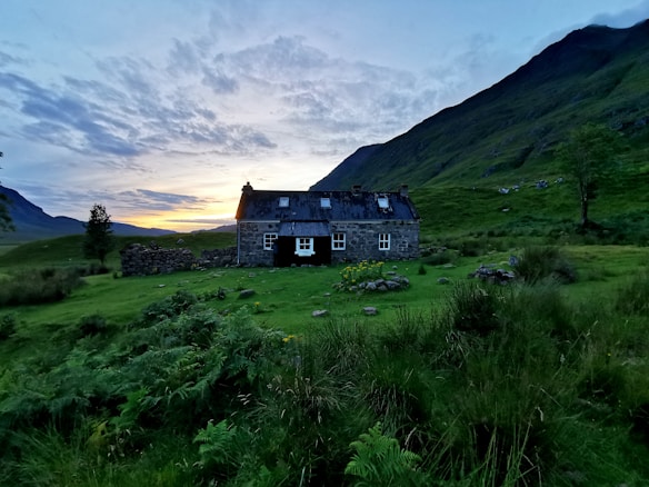 A stone cottage with a dark roof sits in a lush green valley surrounded by hills. The sky is partly covered with clouds and has a warm glow near the horizon, suggesting either sunrise or sunset. There are trees and wild shrubs scattered around the cottage and in the surrounding landscape.
