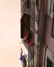 A building facade with clothes hanging on a drying line. A red fabric drapes over a balcony railing, while other garments, including a blue and white piece, hang below. The structure's exterior is visible with a netting or wire fence along the balcony.