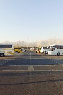 Several parked buses are arranged in a row along a large parking area with visible road markings. People are walking near the buses, and there are mountains in the background under a clear blue sky.