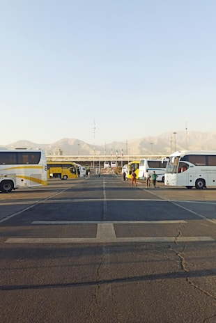 Several parked buses are arranged in a row along a large parking area with visible road markings. People are walking near the buses, and there are mountains in the background under a clear blue sky.