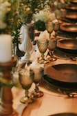 A beautifully set dining table features elegant glassware and tableware. The atmosphere is enhanced by decorative elements like a white candlestick and green foliage entwined along the table. The lighting casts a warm and inviting glow over the entire setting.